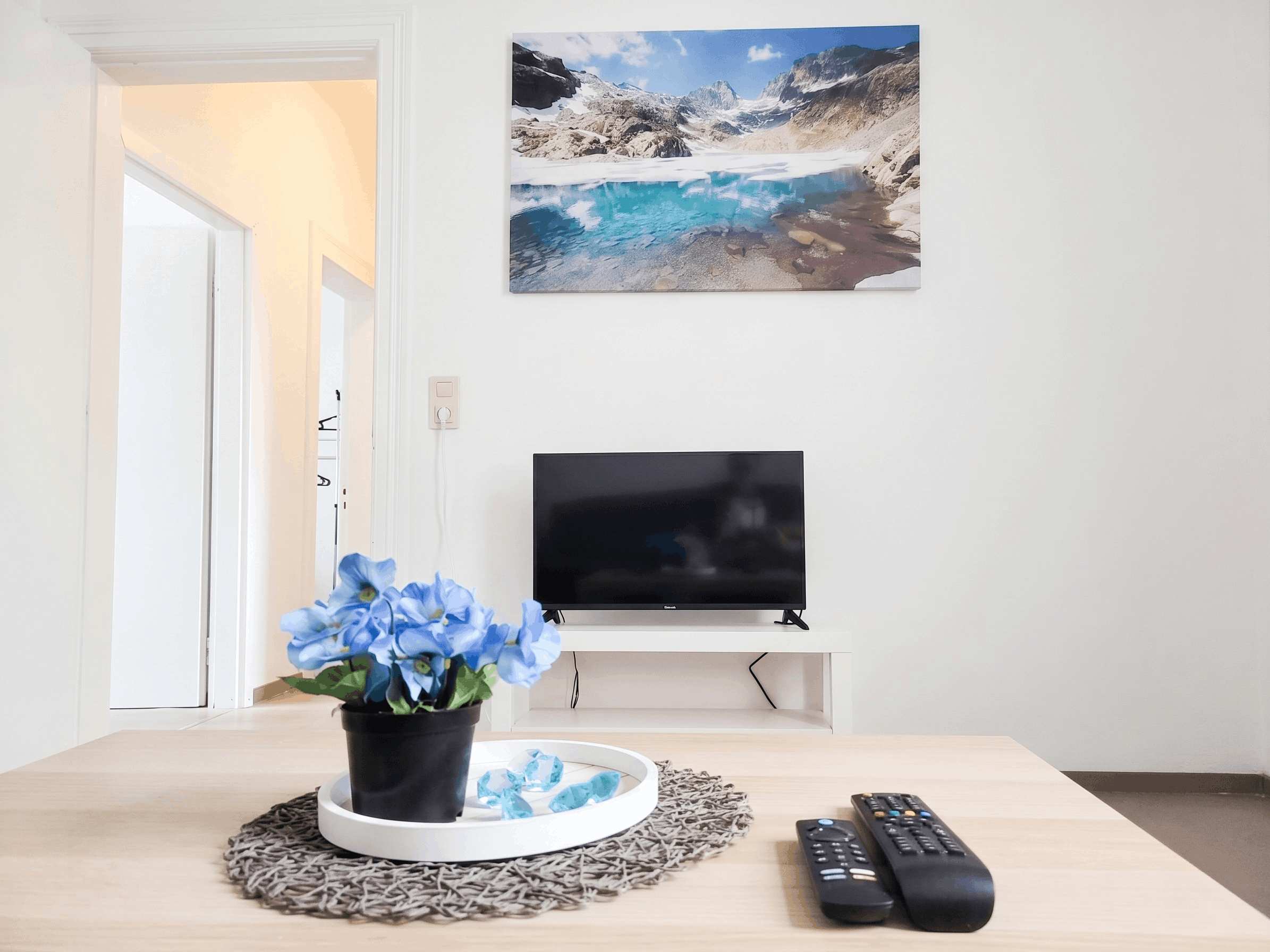 TV – Wohnbereich mit Flachbildfernseher, Couchtisch, blauer Blumendeko und Berglandschaft-Wandbild.